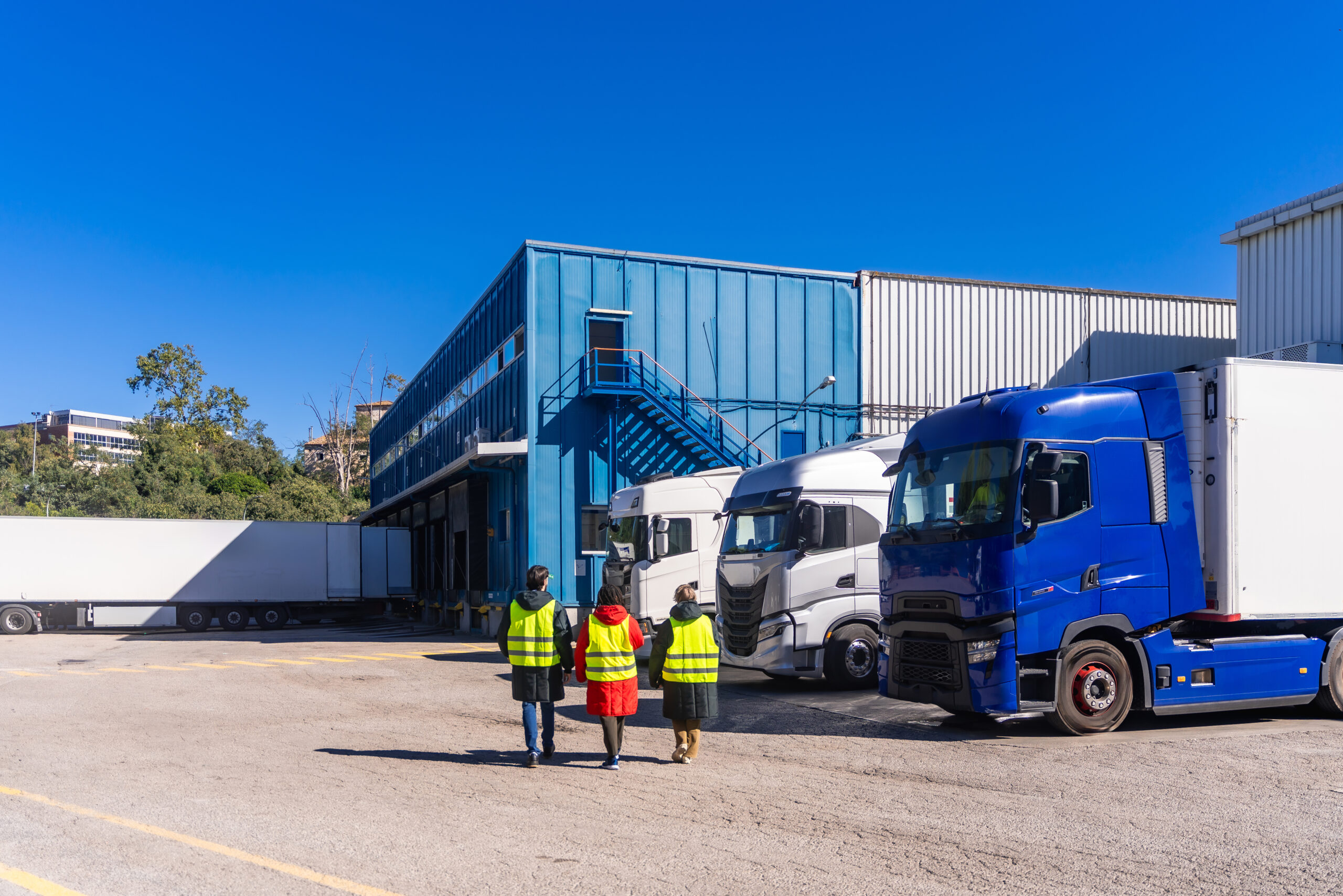 Logistics workers in high visibility vests walking past semi trucks parked at the loading docks of a large industrial warehouse, representing refrigerated transport and cold chain distribution